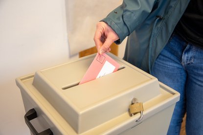 Hand mit Wahlbrief, der schon halb in der Wahlurne steckt